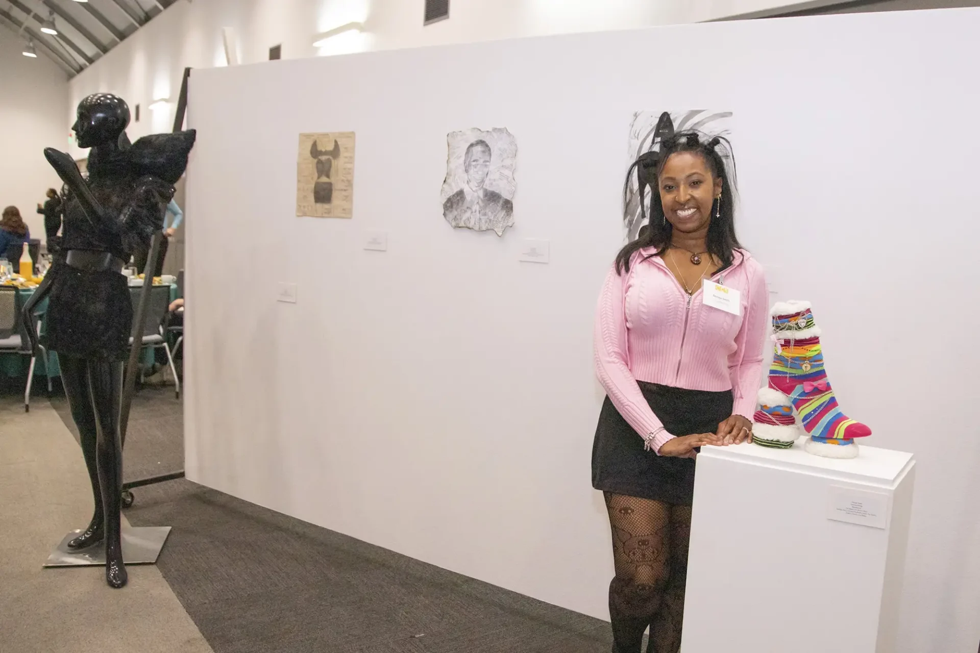 A smiling woman stands in an art gallery setting next to a white display pedestal showcasing a colorful, embellished boot sculpture.