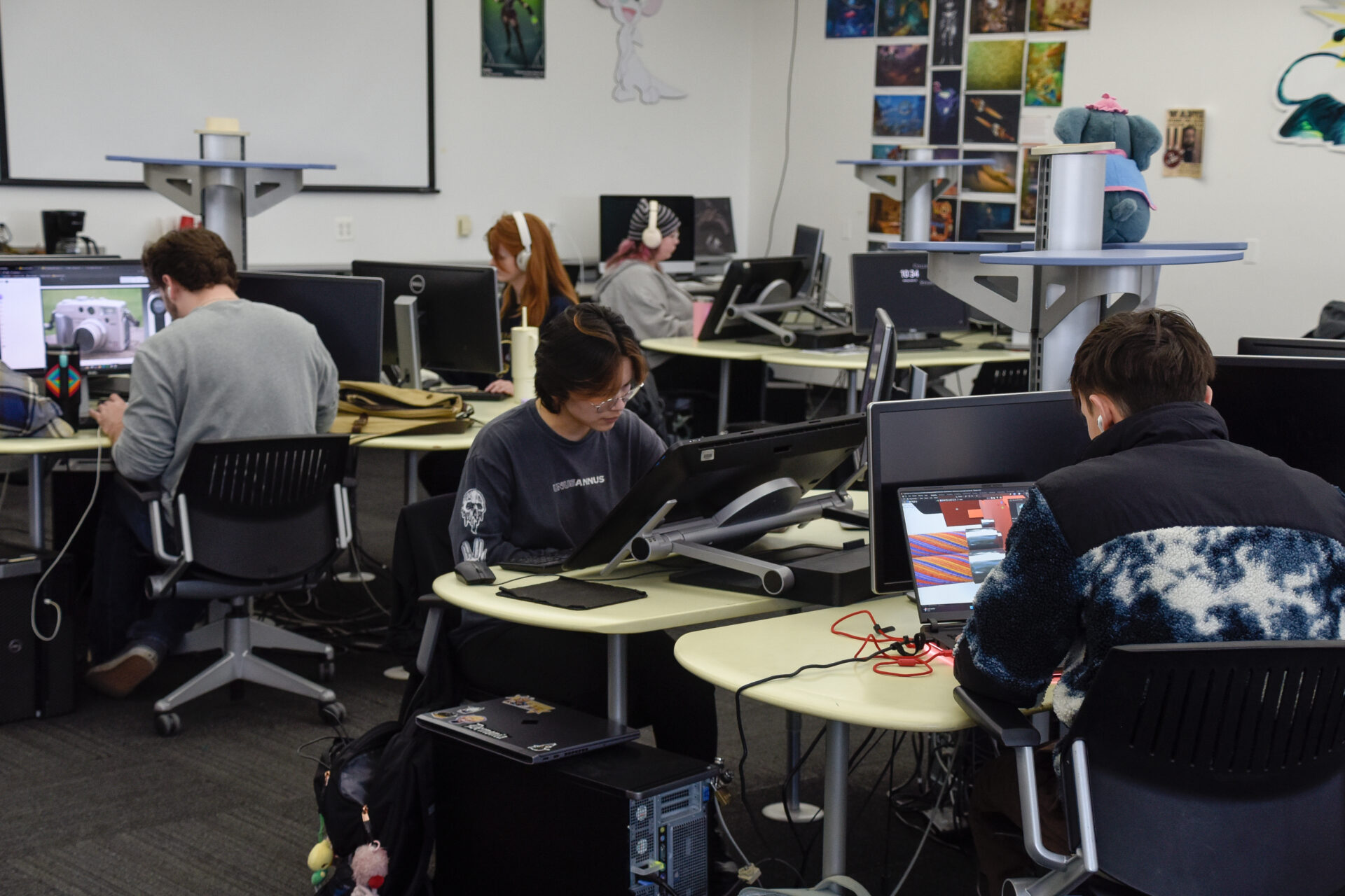 Students work on projects in a game design lab with computers, Wacom tablets and artwork pinned to the wall.