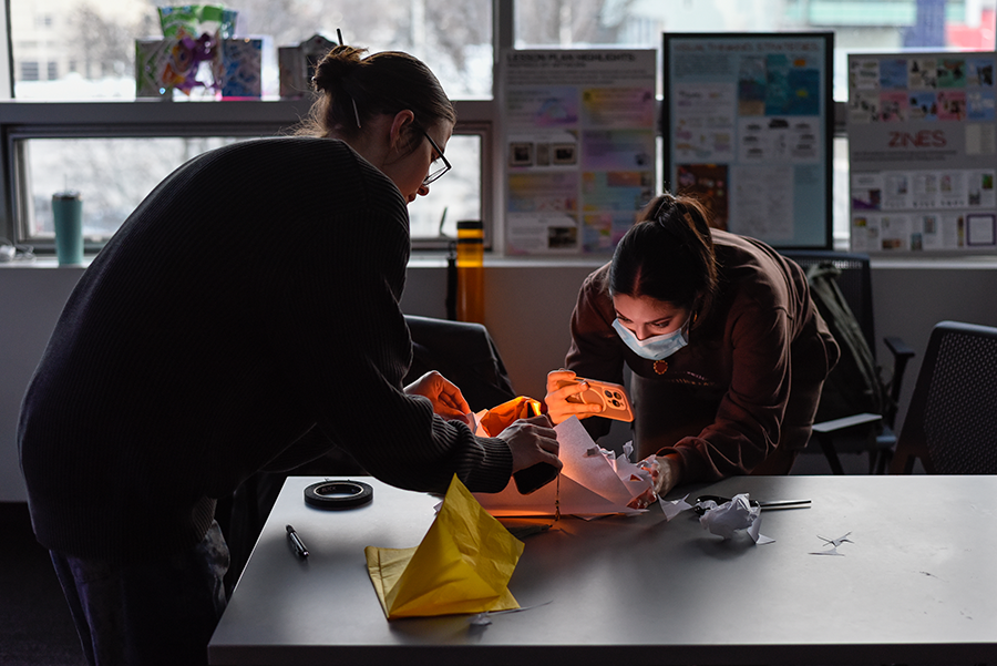 Two students collaborate at a table in a dimly lit classroom, using a smartphone to illuminate and photograph the inside of a handmade paper sculpture, creating a warm orange glow.