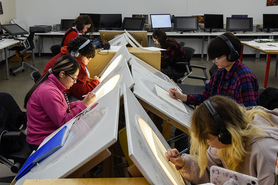 An interior view of a busy animation studio classroom. In the foreground and midground, multiple students wearing headphones are seated at long rows of angled drafting tables. Each table features an illuminated circular light box used for traditional 2D animation sketching. The students, including one in a bright pink jacket and another in a plaid shirt, are intently focused on drawing. In the background, rows of desktop computers and digital drawing tablets line the wall of the well-lit room.