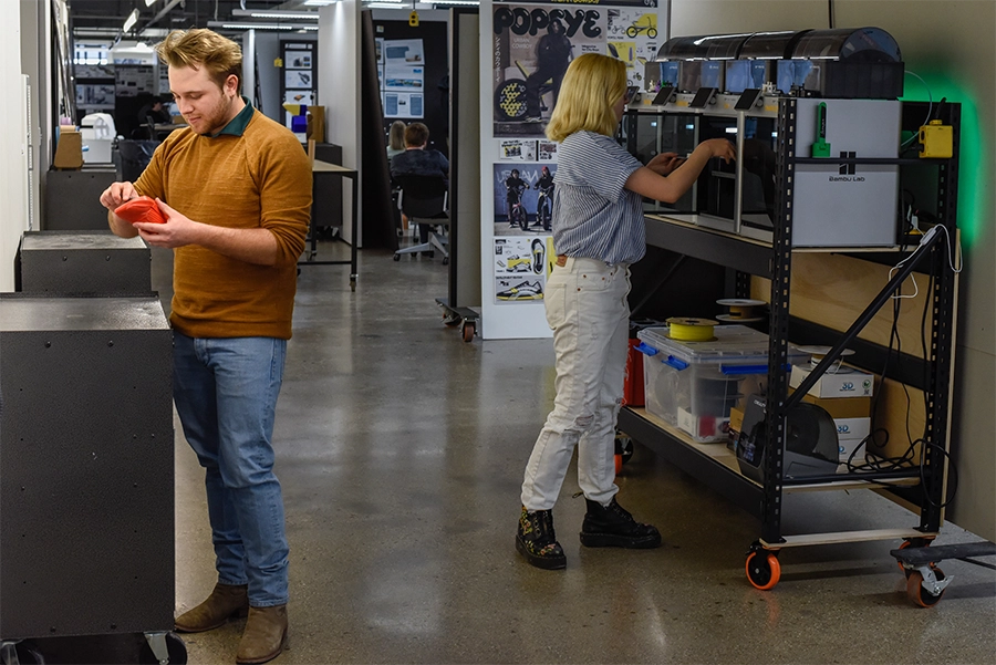Two students working in the Product Design space, one working with 3D printers and the other looking at a red 3D printed shoe.