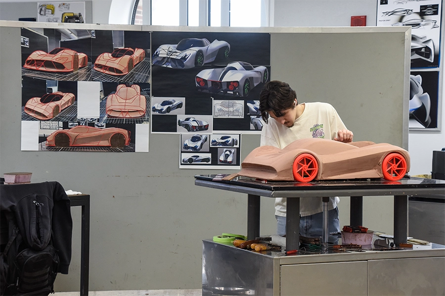 A student works on a clay model of a car with orange wheels in the CCS Transportation Design studio with artwork hanging on the wall in the background.