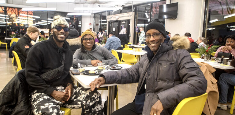 Three smiling people sitting around a white table in a bright indoor room with large windows.