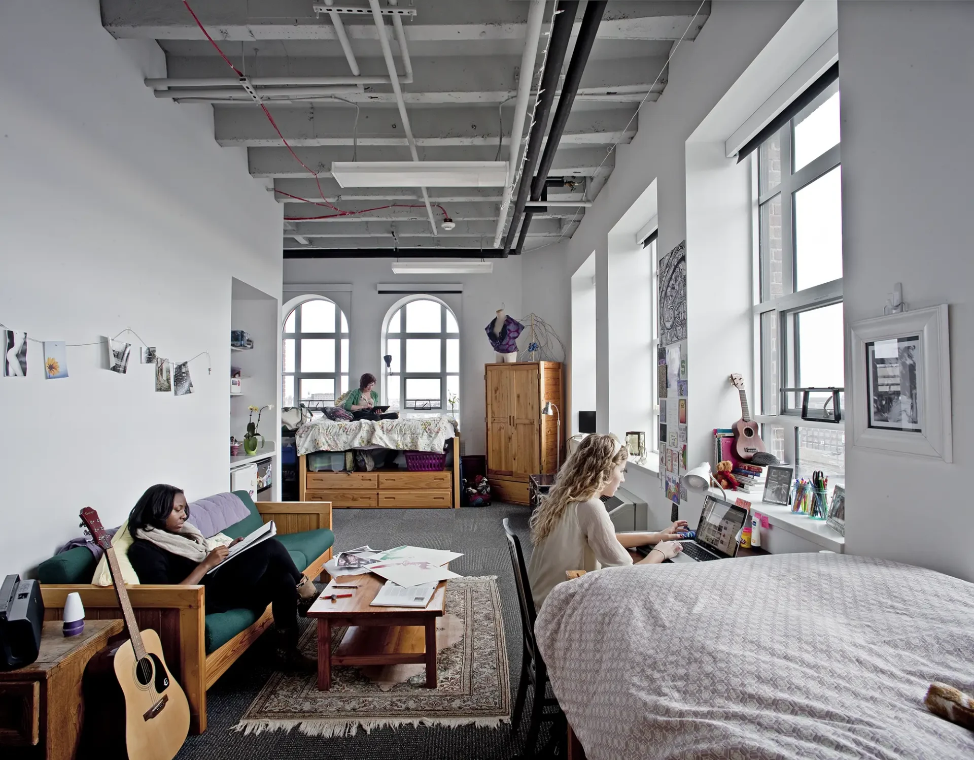 Students lounging and working in a large open dorm space. There are large sunny windows