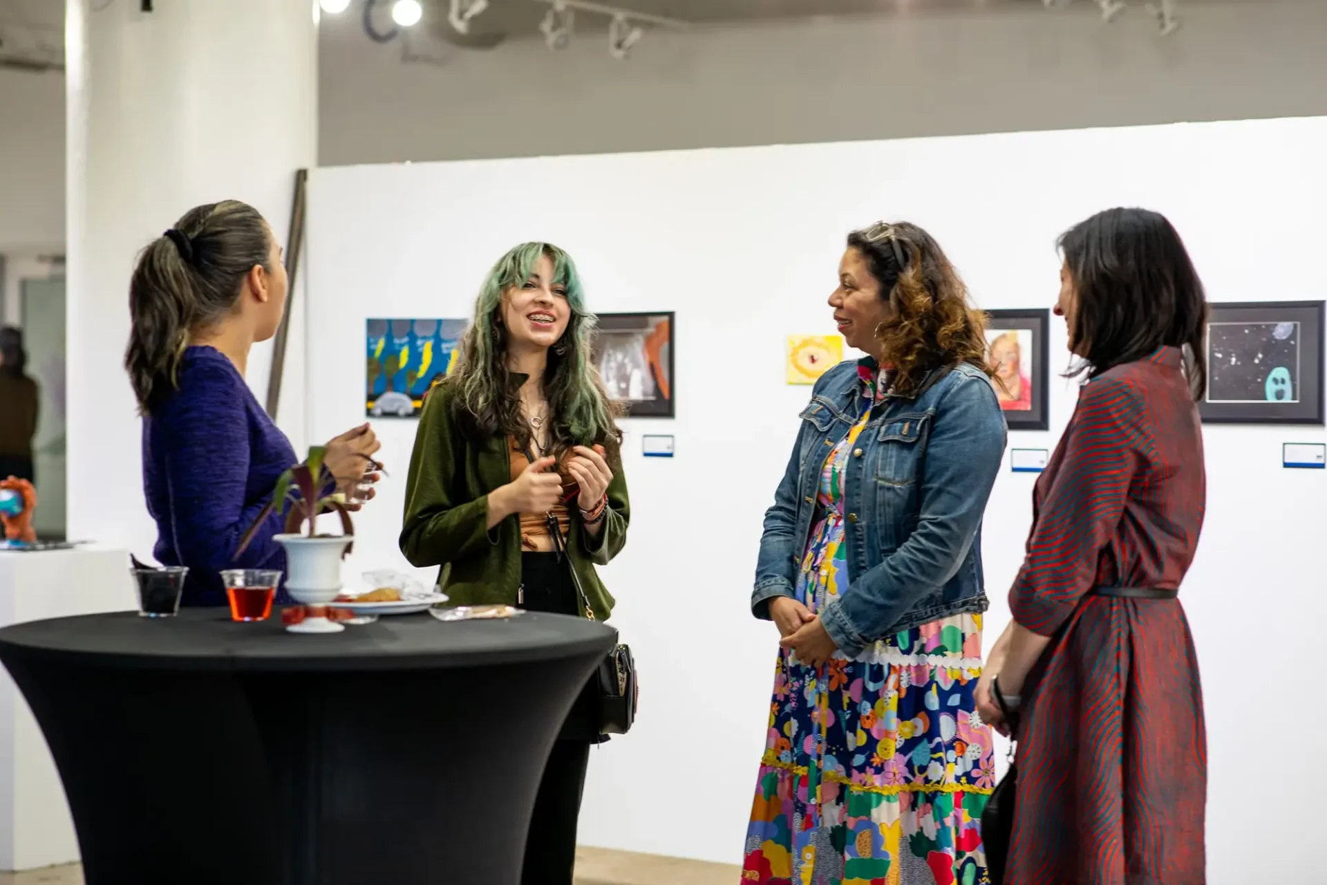A group of women talking and laughing with drinks around a small table in a gallery space