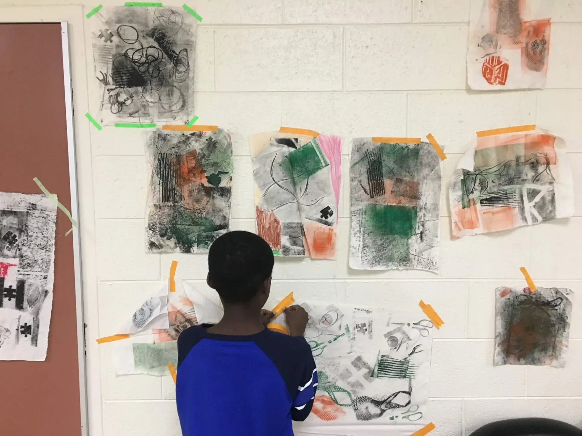 A young boy taping a piece of artwork to a wall covered in drawings and paintings