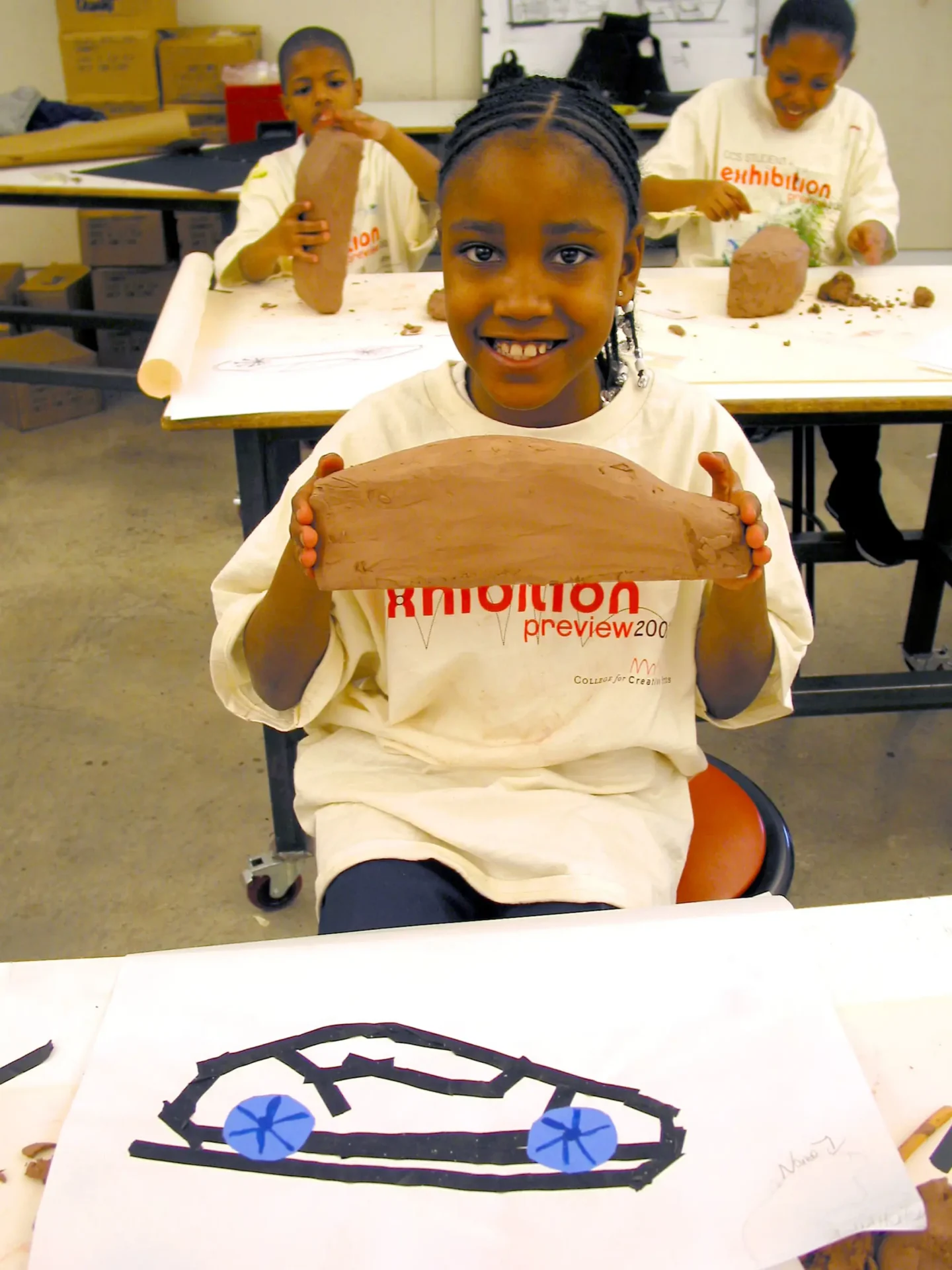 A little girl holding up a clay model of a car she sketched and smiling