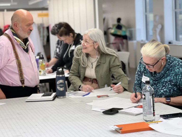 Three people gathered around a table. Two are speaking, one is sketching something