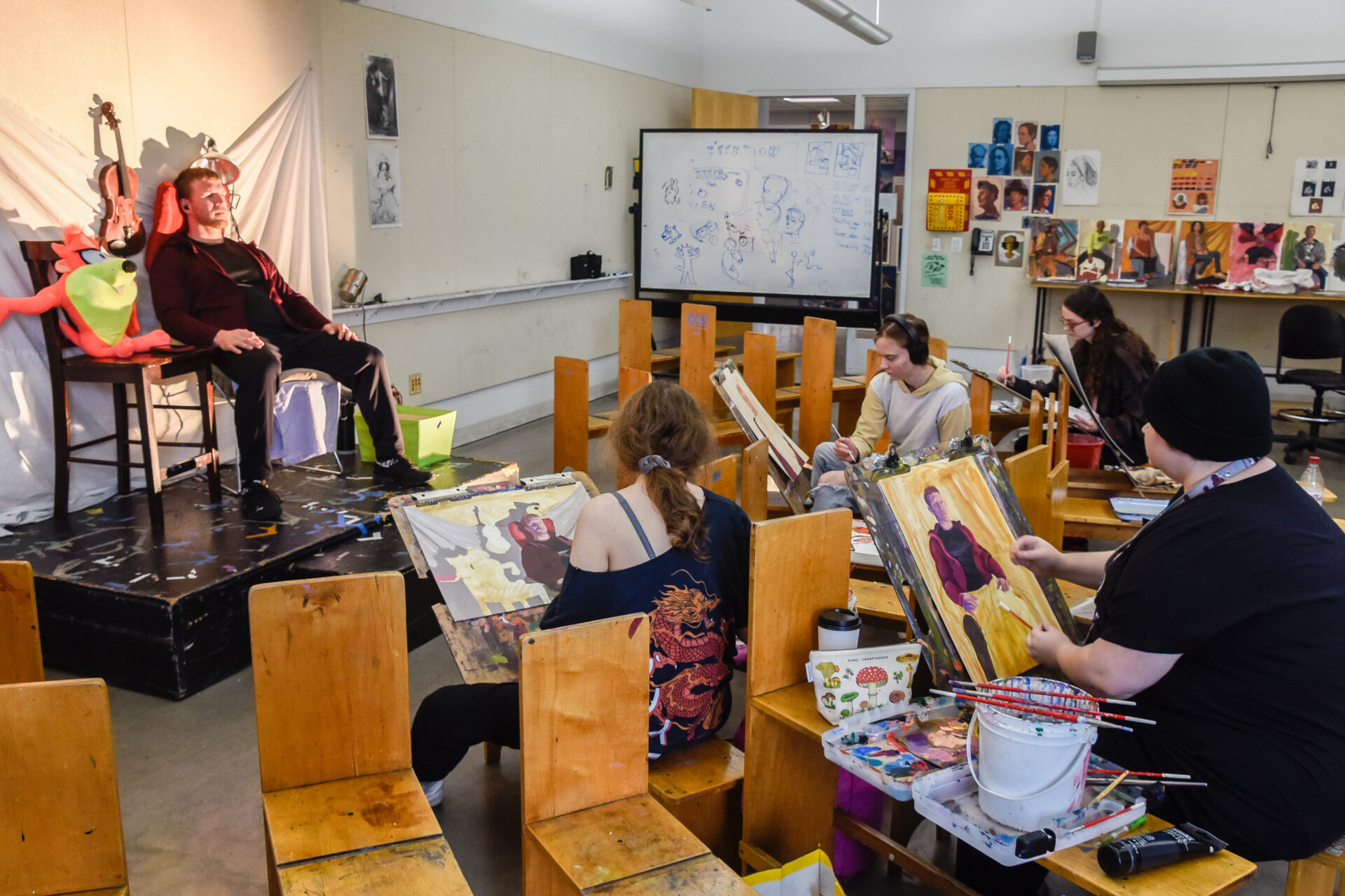 An art classroom setting where a live model sits on a small stage at the front left, posing for a group of students who are painting him. The model, a man in a maroon jacket and dark pants, is seated with a neutral expression. Behind him on the stage, a violin hangs against a white draped backdrop, and a bright orange-and-green cartoonish plush character is draped over a chair next to him. In the foreground and midground, several students sit on wooden chairs facing the stage, working on their paintings. On the right, a student in a black beanie and shirt works on a canvas, applying yellow and maroon paint that depicts the model. Another student with long brown hair tied back wears a dark shirt with a dragon design and looks toward the model. In the background, a large whiteboard filled with sketches and diagrams stands near the center, and the back wall features a display of various student portraits and artwork.