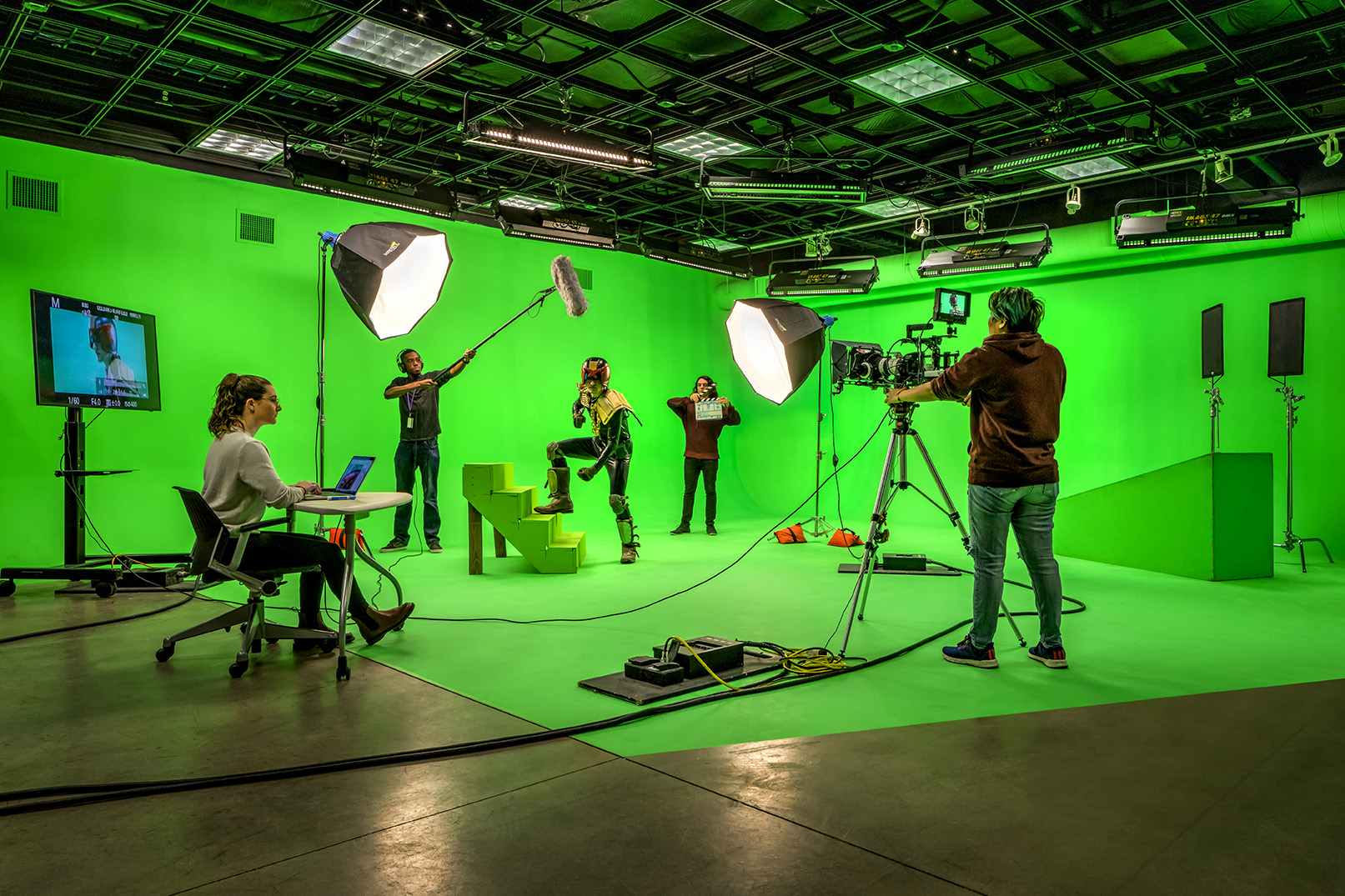 A production crew and actors working inside a professional, fully equipped green screen studio at CCS. A camera operator films a costumed actor standing on green steps, while a boom mic operator captures audio and another crew member holds a clapperboard. A student sits at a desk in the foreground monitoring the live footage on a laptop and nearby display.