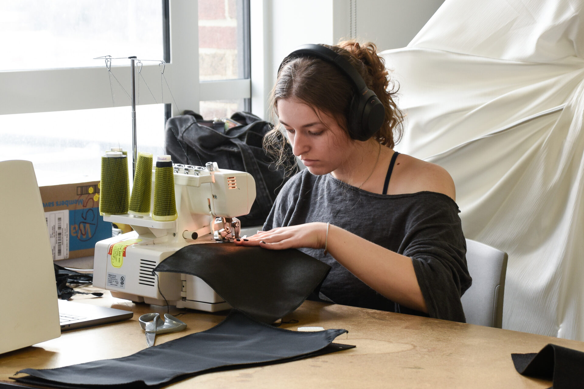 A student sits by a window at an industrial sewing machine sewing a piece of black fabric with a white garment draped in the background.
