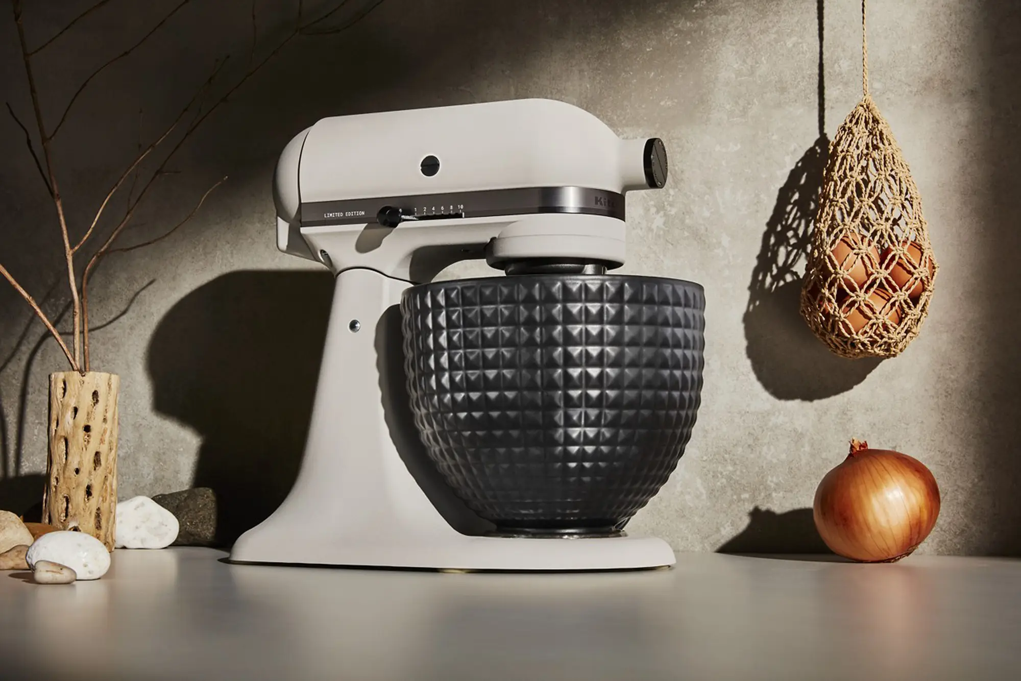 A stand mixer on a counter top, next to some decorations, and a sack of onions hanging down. The image is dramatically lit