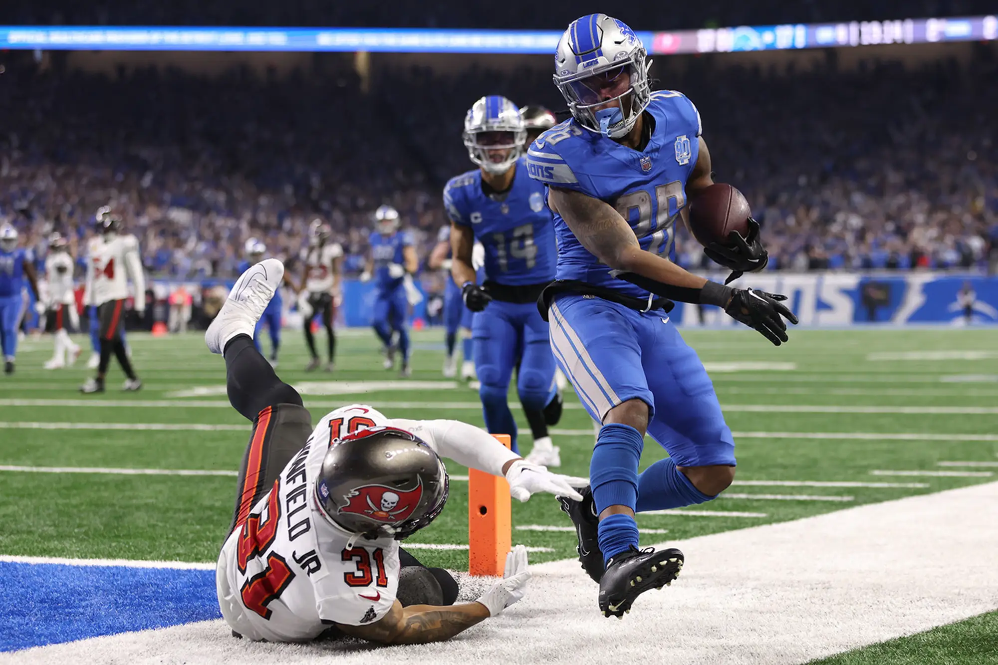 A Lions player scoring a touchdown, stepping over a defender from the Tampa Bay Buccaneers at Ford Field