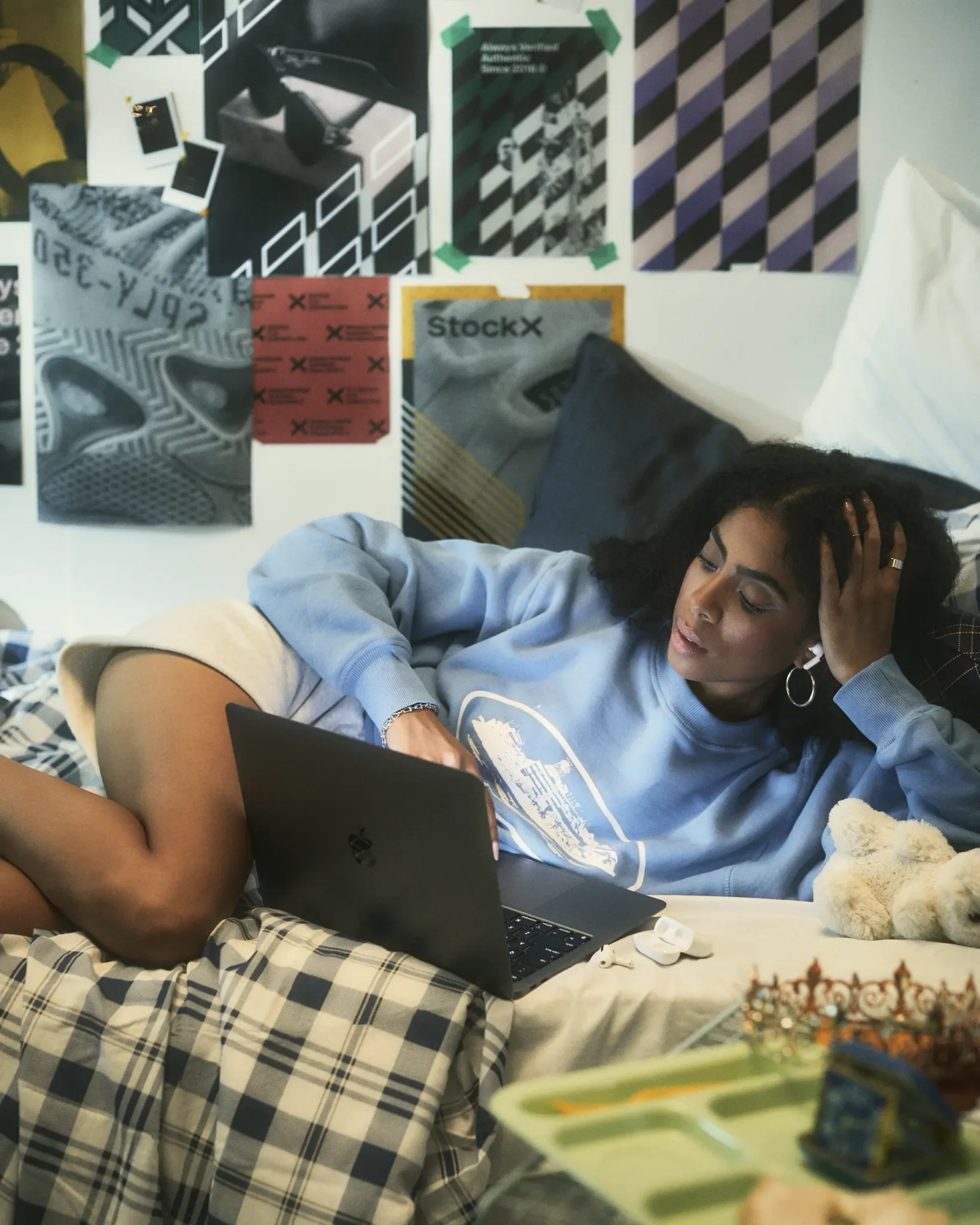 A young woman on a bed, browsing on her laptop. Behind her is a wall filled with posters and images branded with StockX