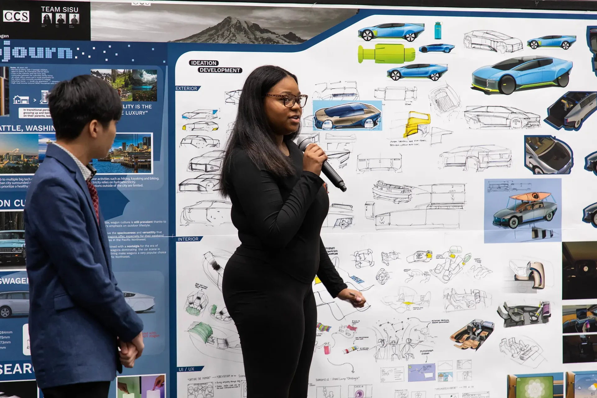 Two students speaking into a microphone in front of a board full of auto sketches and renderings