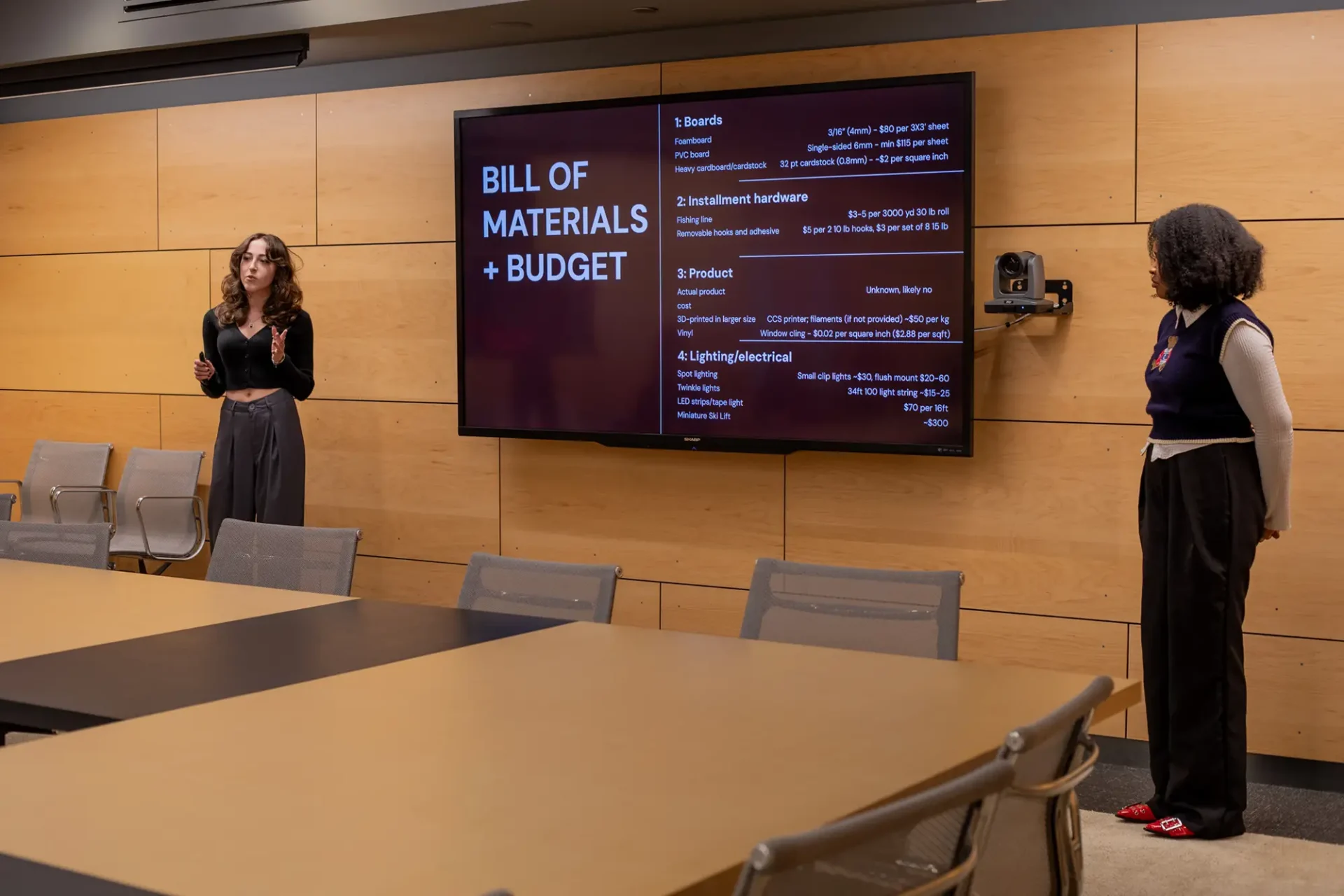 Two students presenting on a large screen about Bill of Materials and budget