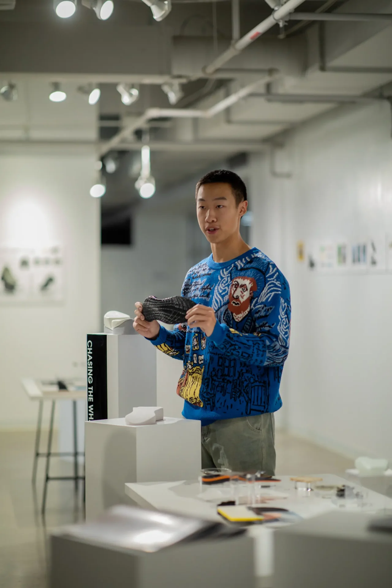 A student holding up a shoe sole, presenting his work. Multiple small white podiums around him feature materials