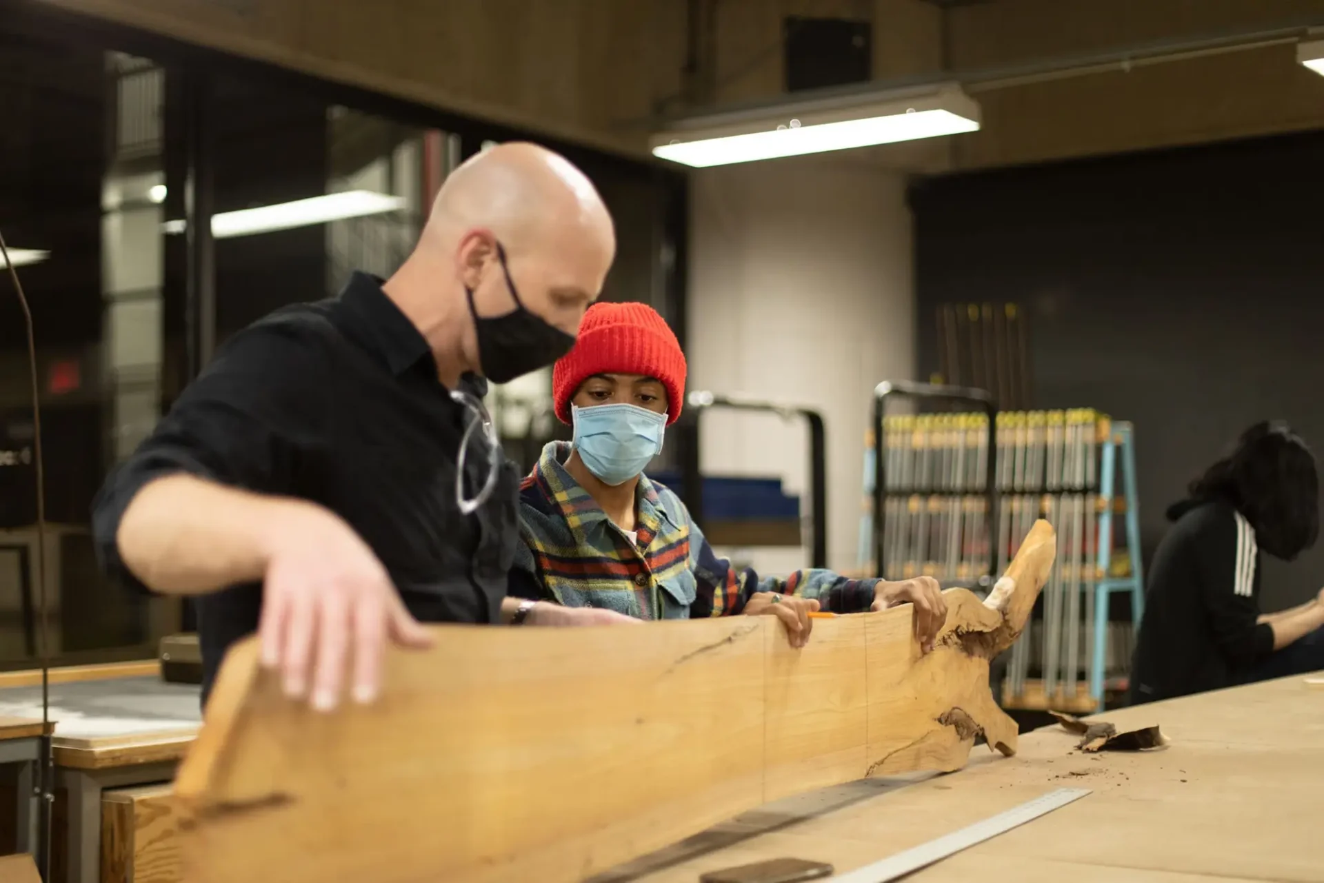 Two people holding up and looking at and measuring a large plank of wood at a table
