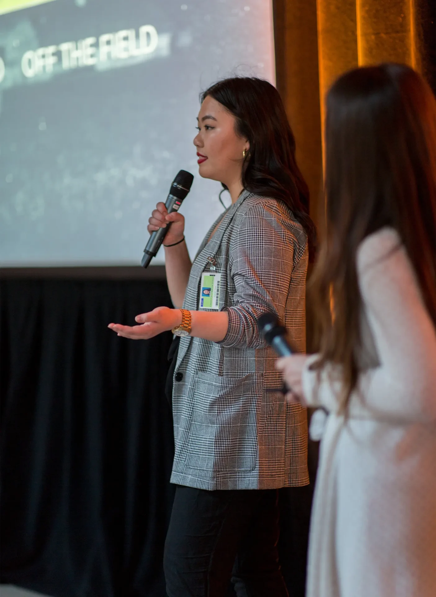 A woman presenting, holding a microphone next to another woman, also with a microphone