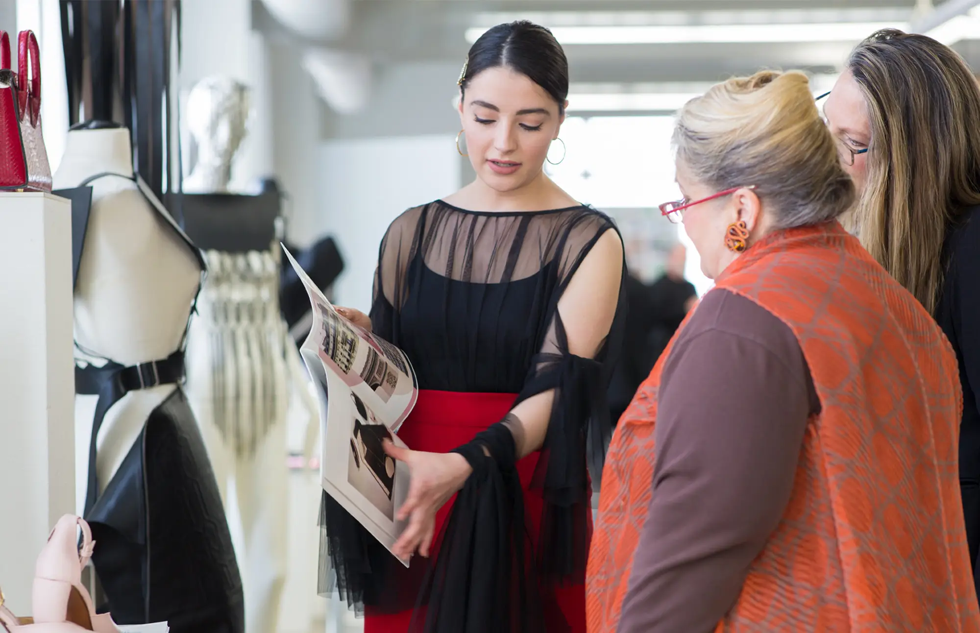 A woman showing a look book to two other women, next to a mannequin bust wearing a black dress