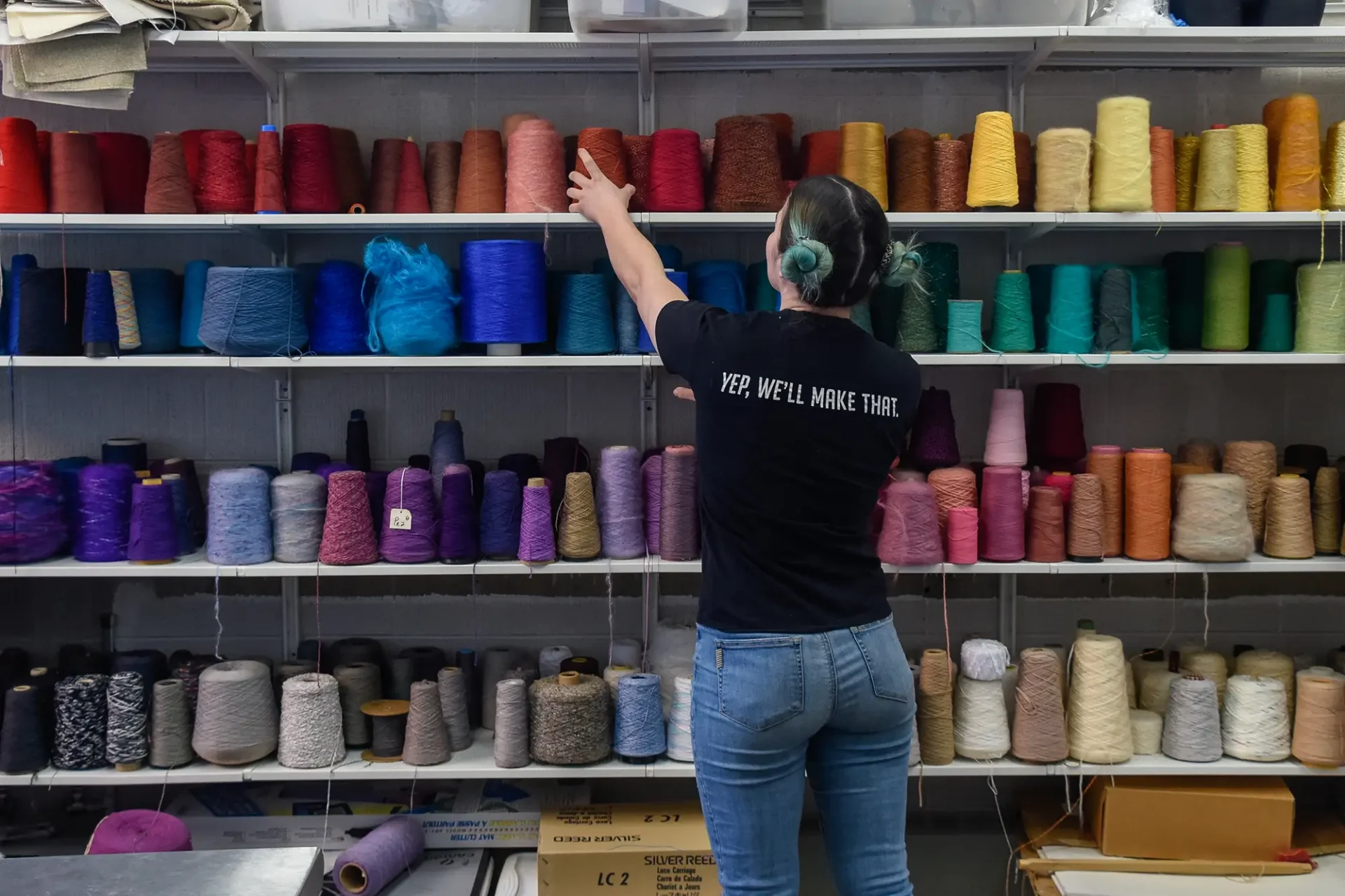 A student reaching for a spool of yarn on a shelf full of large spools of colorful yarn