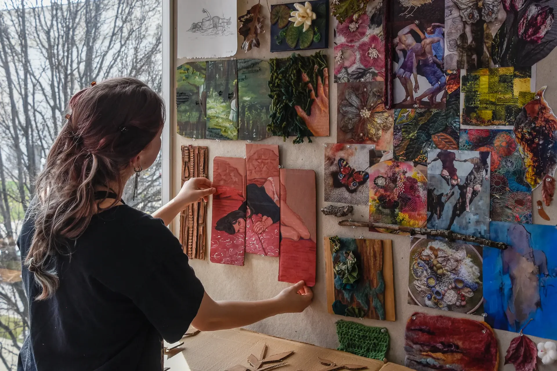 A student pinning a tryptic painting onto a board full of drawings and paintings next to a well lit window