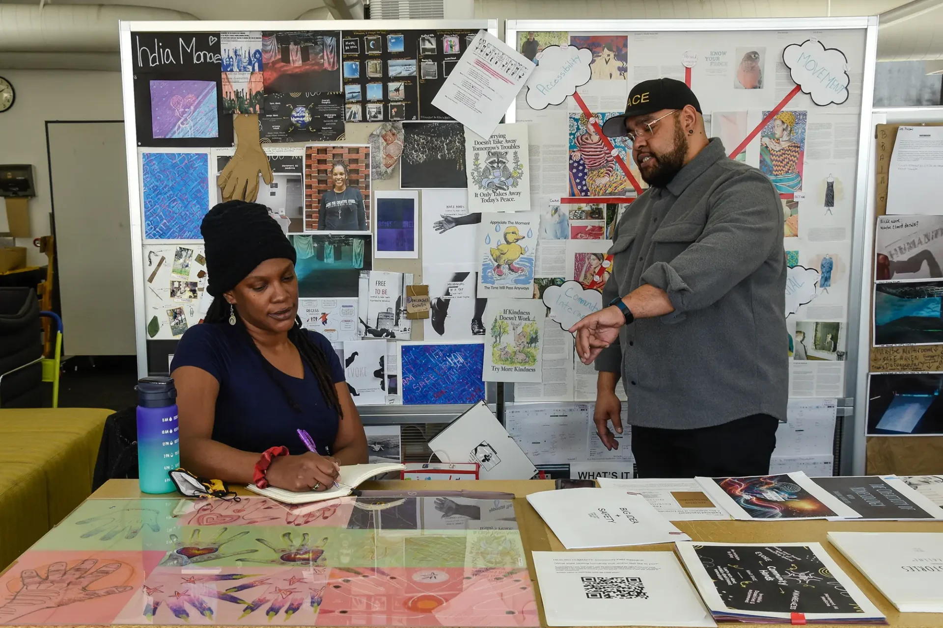 Two students working at a table. Behind them is a large mood board fill of images, writing and inspiration.