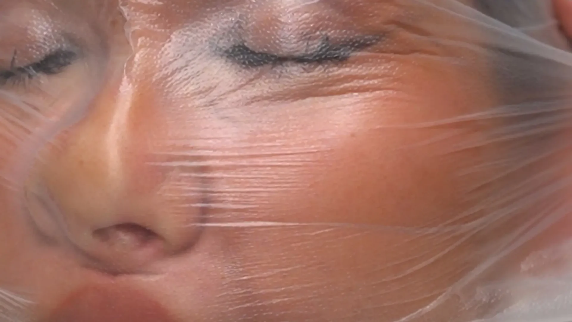 Film still from performance short: extreme close-up of female performer's face wrapped in plastic.