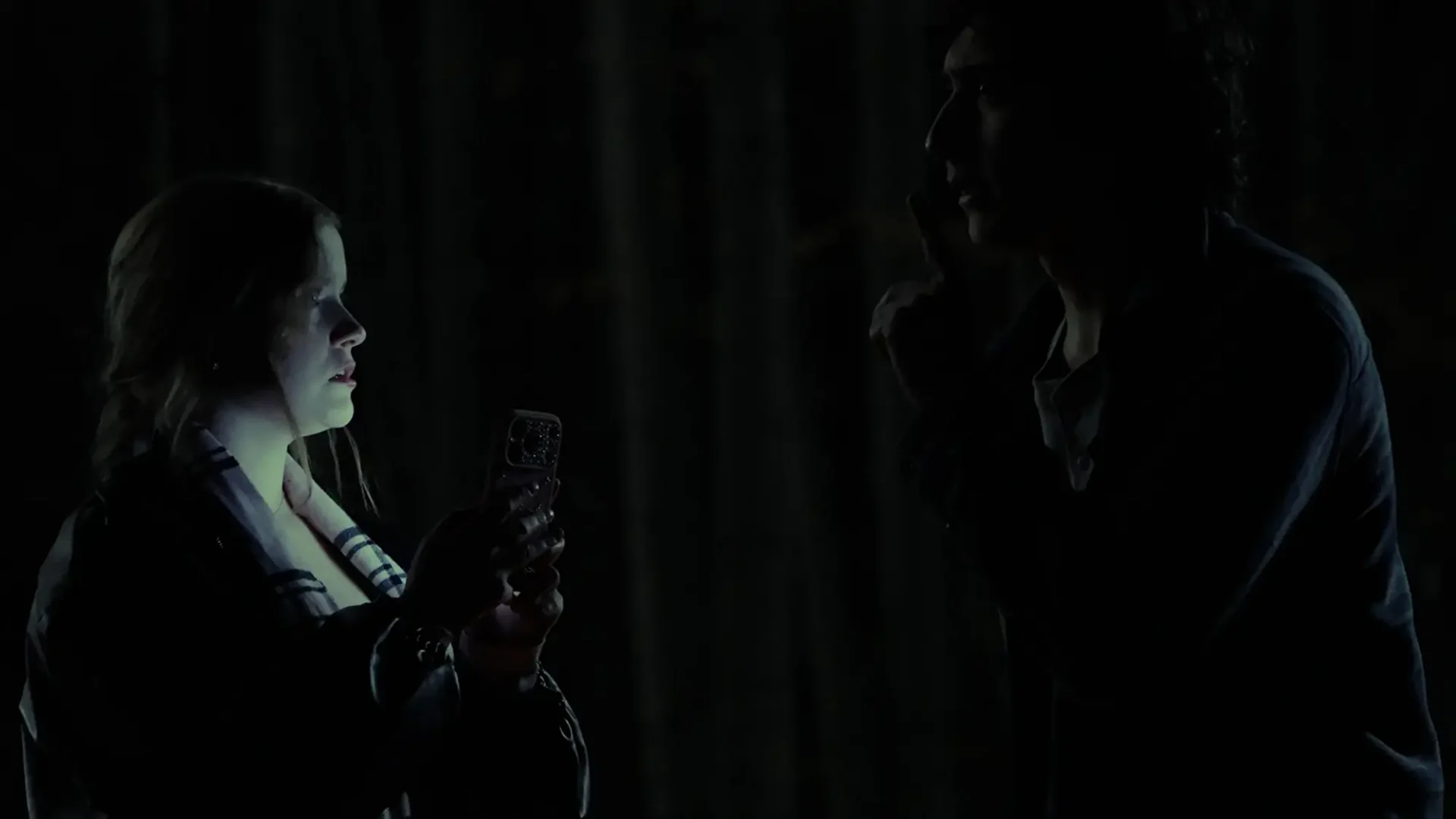 Two shot of a woman and man facing each other in the dark, she is illuminated by her cell phone.