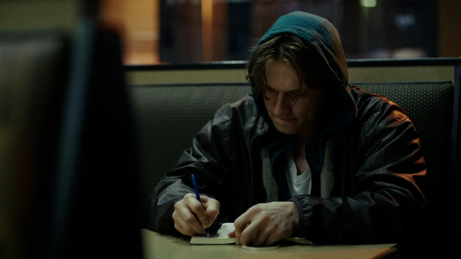 Film still from 2K thriller Random Bathrooms: Medium shot of a young man sitting a booth at a diner scribbling a notebook.