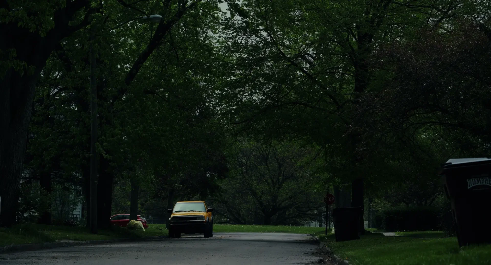 Film still: a suburban street in Fostoria, OH with a car parked at the end of the block near some trash on the curb.