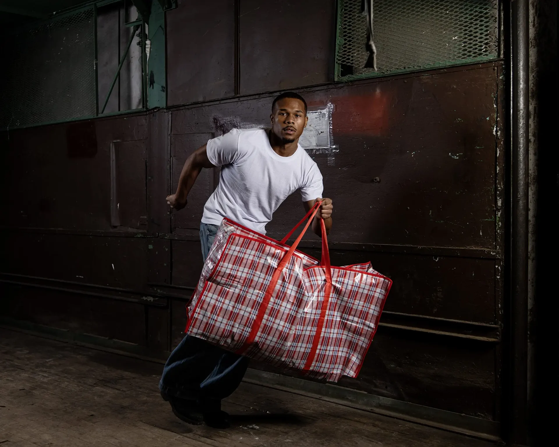 A model runs through an industrial hallway carrying a plaid laundry bag starign directly into the camera.
