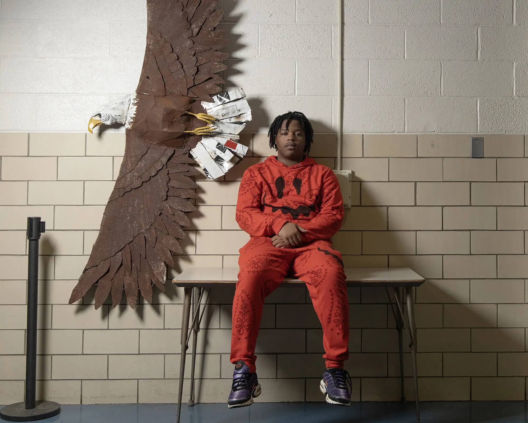 Environmental portrait of a tween-age boy sitting on a table in a school hallway with a large papier mache eagle on the wall.