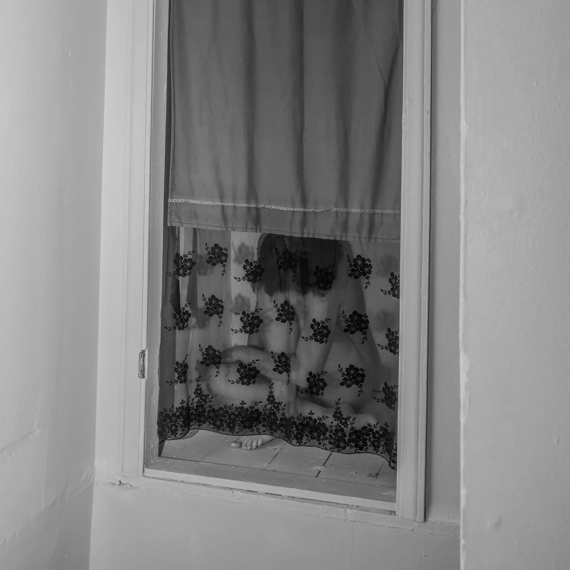 A girl hides behind a sheer curtain in the corner of a room.