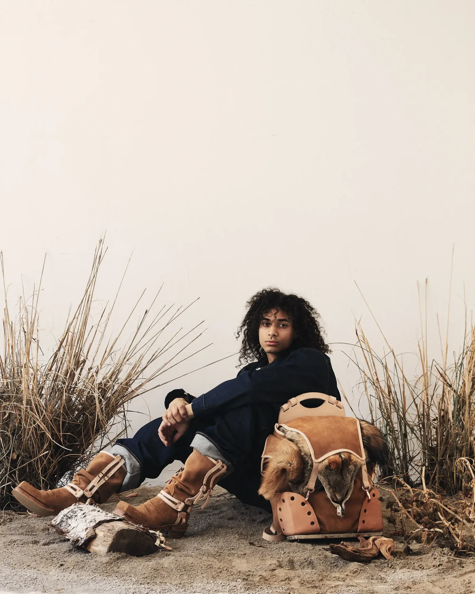 Model wearing leather boots sitting next to leather backpack with animal hide