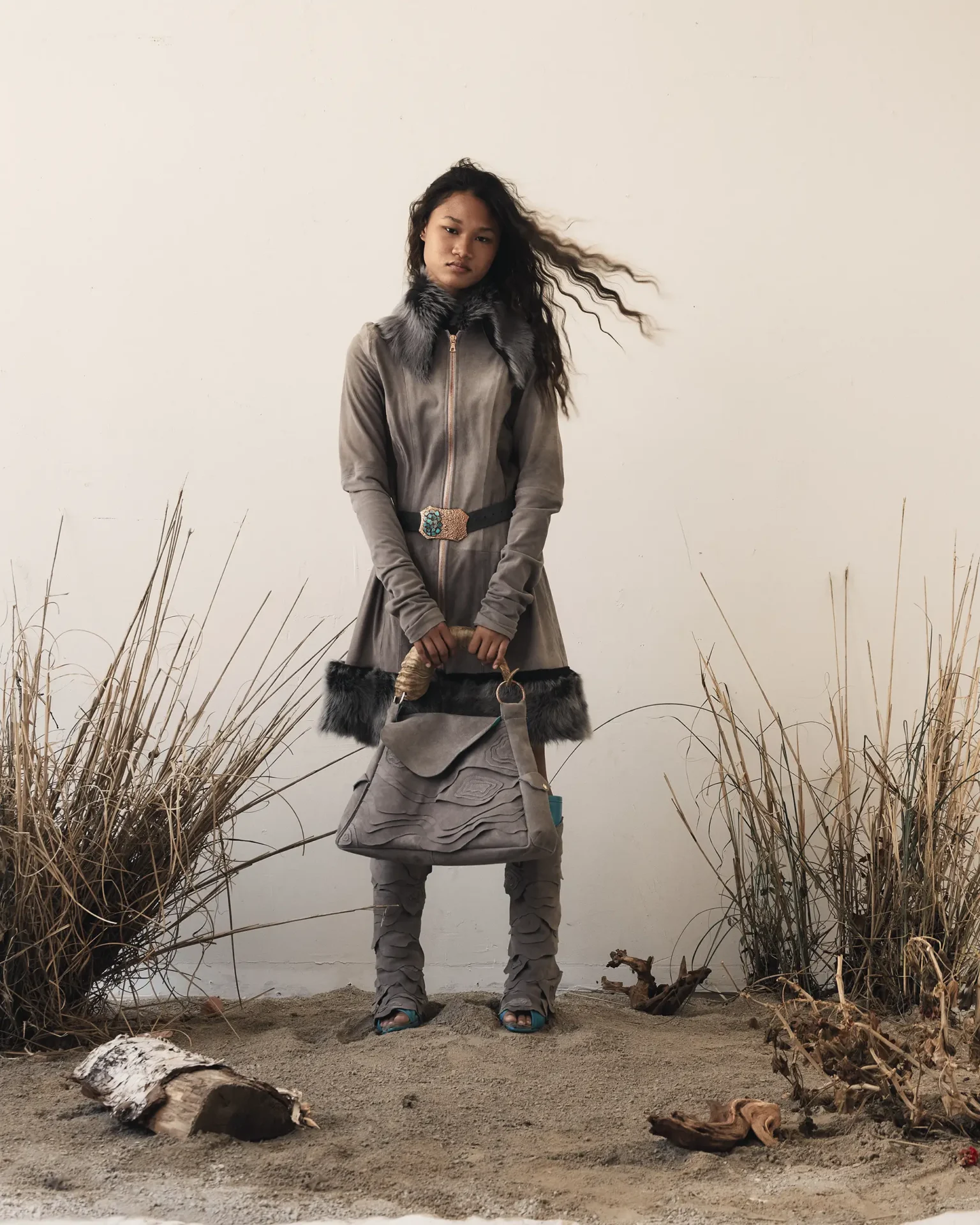 Model wearing suede coat with fur collar, holding bag with horn handle and wearing knee high boots