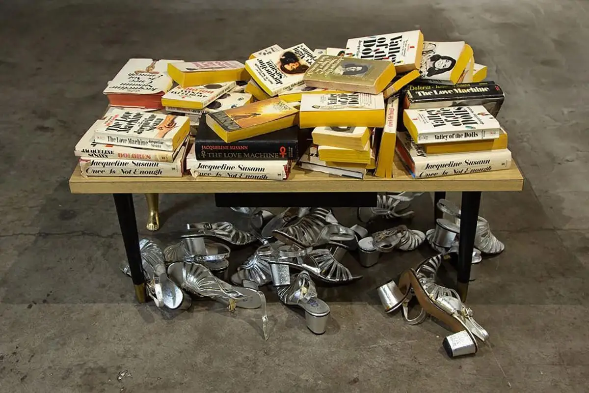 A table, covered in messily stacked books with a pile of silver women's shoes scattered beneath it