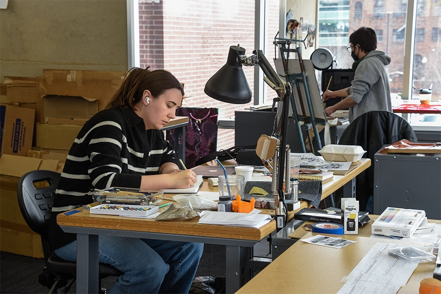 Students working in a studio with wide open windows, large desks and a variety of materials, boxes in the foreground and easels in the background.