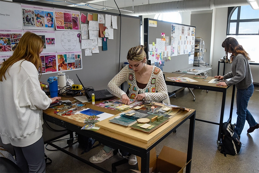 Interior design students work on material boards at large tables in the classroom next to walls filled with design inspiration.