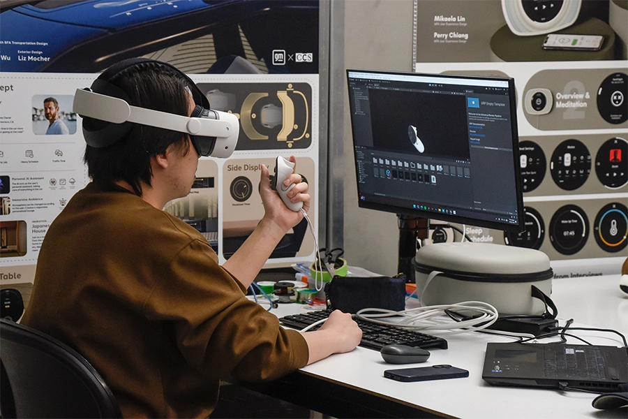 A student sits at a desk in front of a computer monitor with a VR headset on and artwork in the background