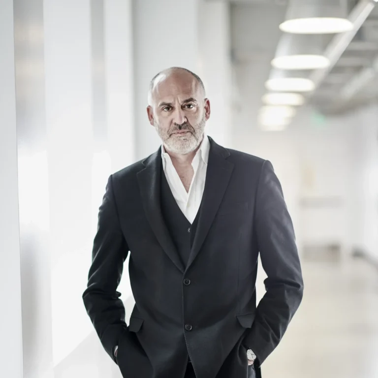 A headshot of Paul Snyder, a mature man where a black suit and white button up shirt standing in a white hallway.