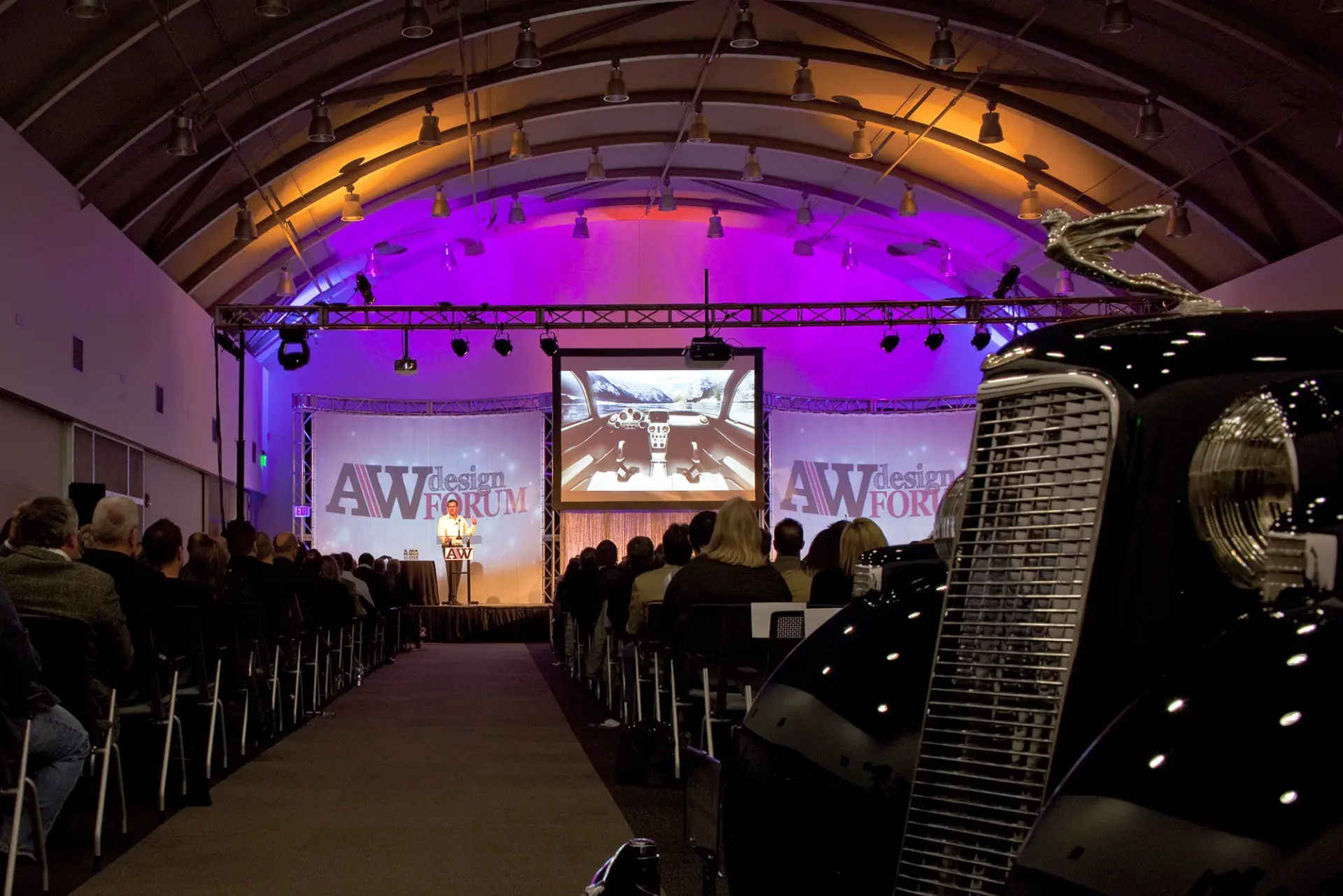 An audience watches a presentation at the AW Design Forum under a purple-lit arched ceiling. The chrome grille and hood ornament of a vintage black car are close up in the foreground.