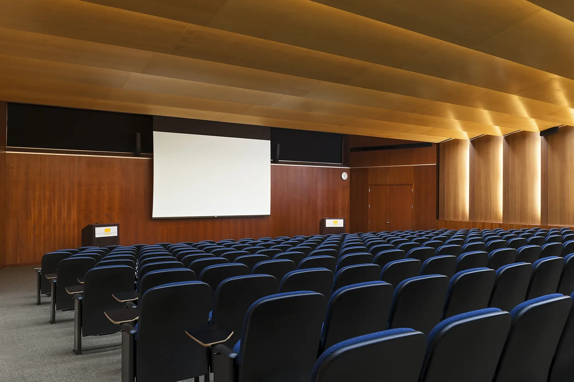 An empty modern lecture hall or auditorium with blue seats and a large presentation screen.