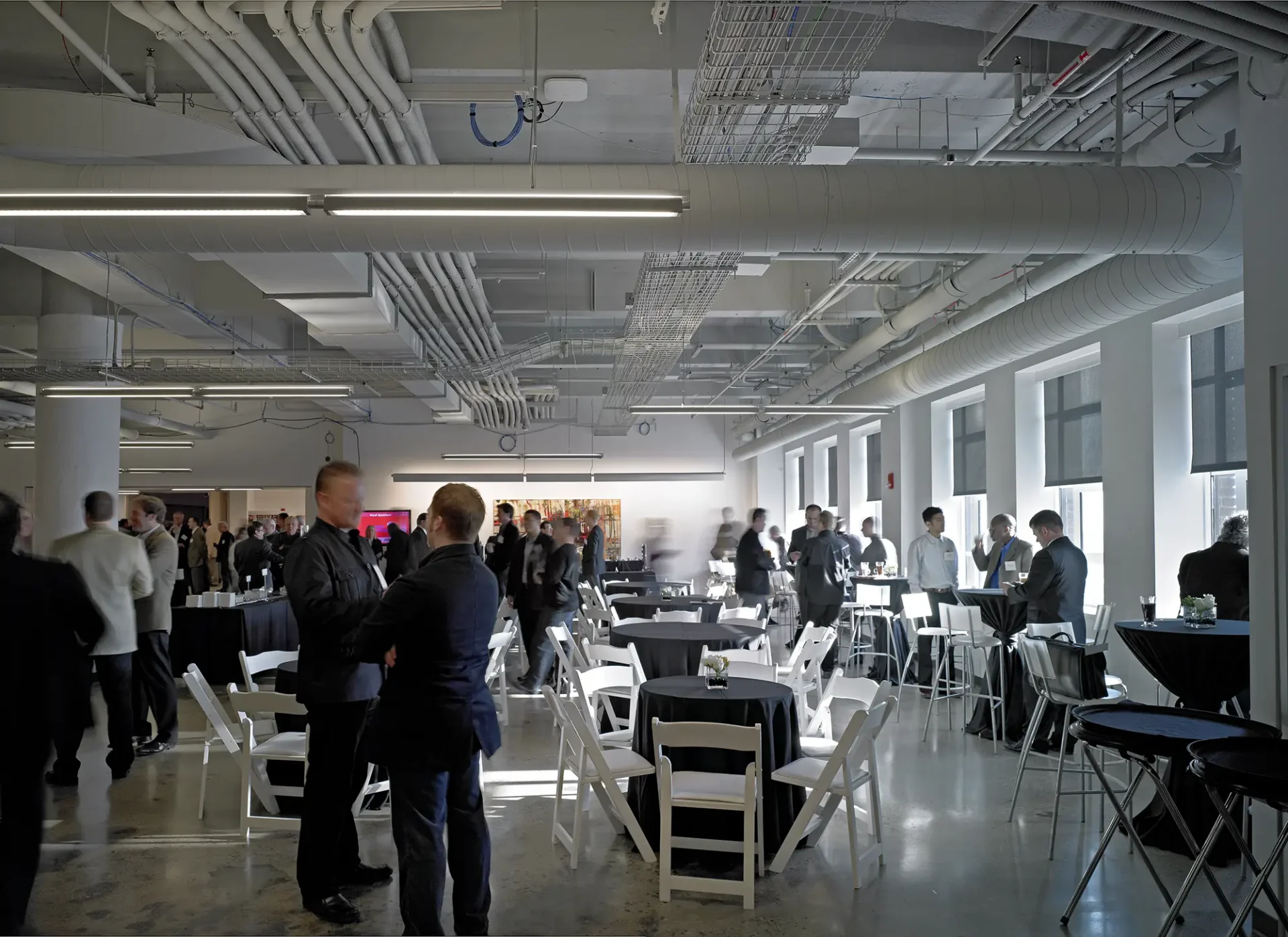 A crowded indoor event in a large room with a polished concrete floor and an exposed ceiling showing pipes and ducts. People are mingling around small round tables covered in black tablecloths with white folding chairs.