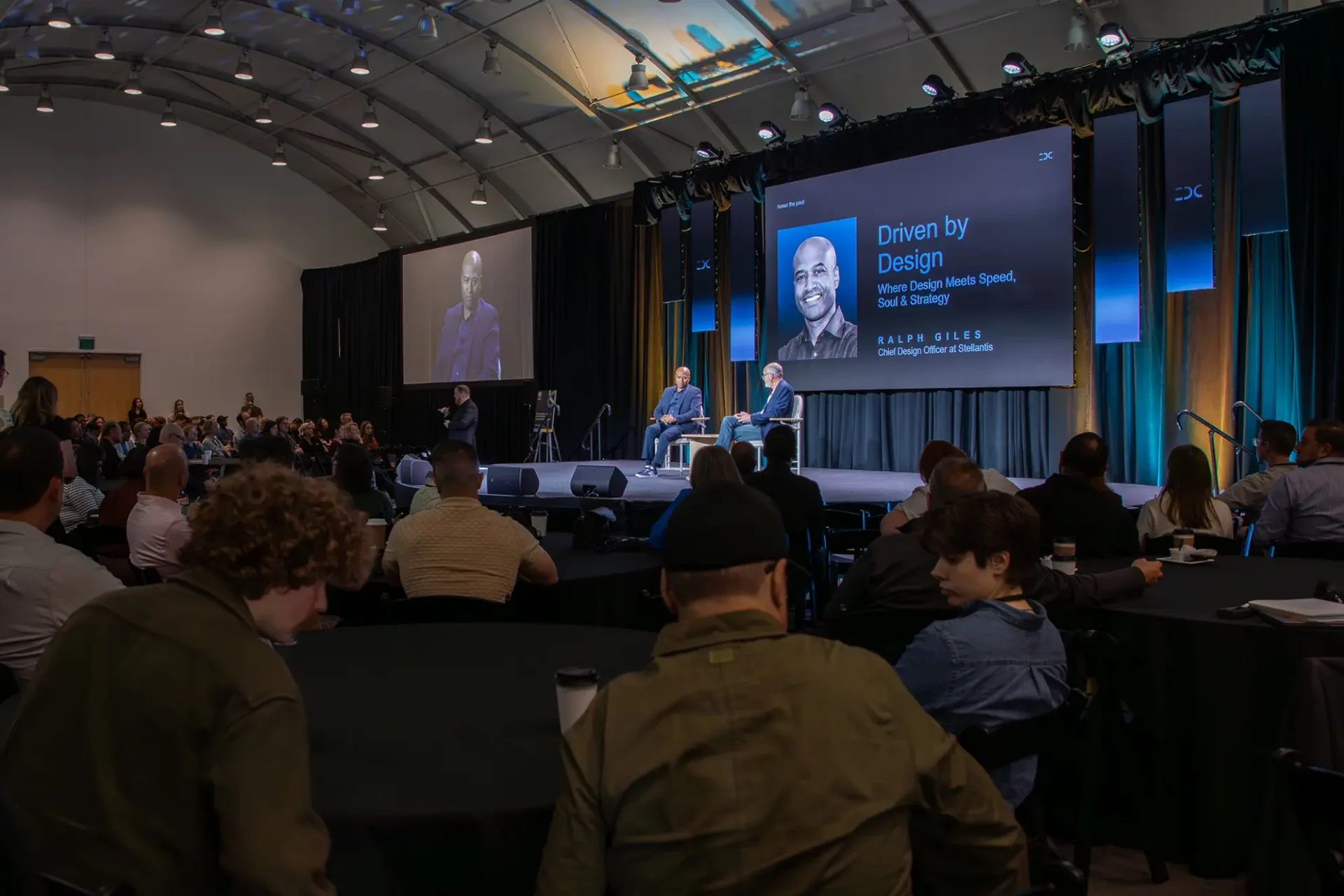 An auditorium presentation with two speakers on a stage beneath a large screen reading "Driven by Design" featuring Ralph Gilles. An audience sits at round tables under a high, arched ceiling.