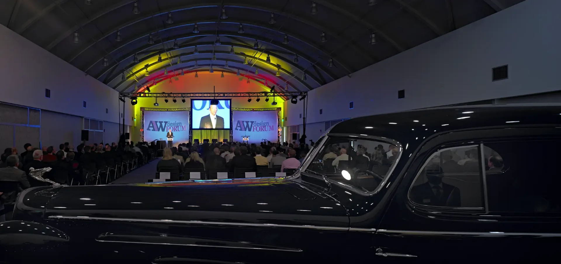 A wide view of a crowded conference hall with colorful rainbow ceiling lights during a presentation, partially framed by the long, glossy silhouette of a vintage black car in the foreground.