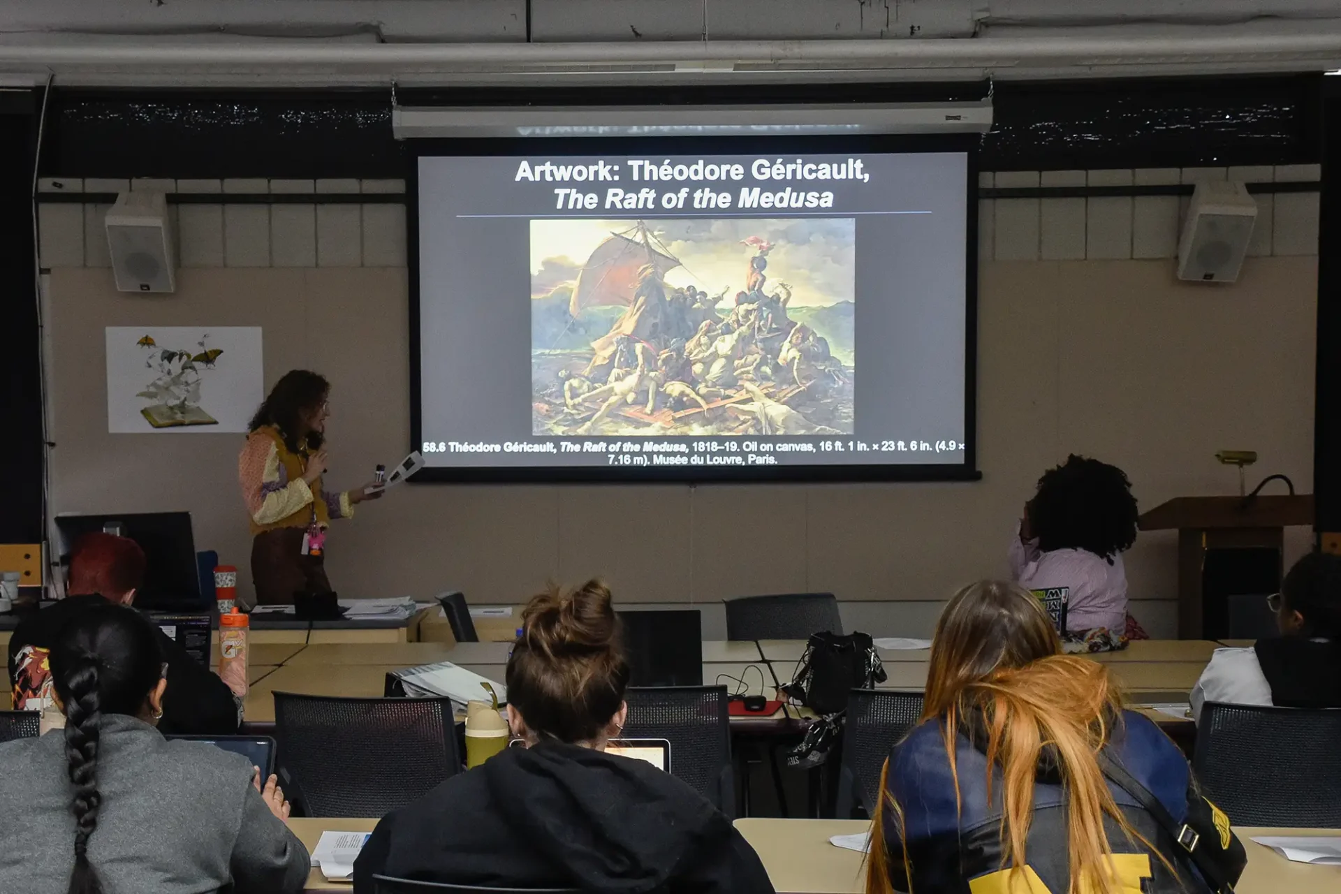 An instructor stands at the front of a dimly lit lecture hall, presenting a slideshow about Théodore Géricault's "The Raft of the Medusa" to a class.