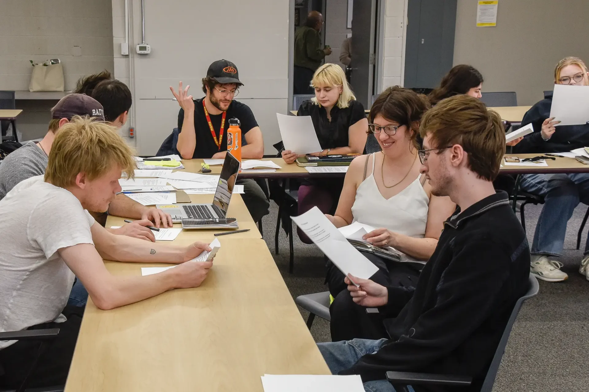 A group of university students are gathered around long tables in a classroom, actively discussing, reading printed documents, and working on laptops together.