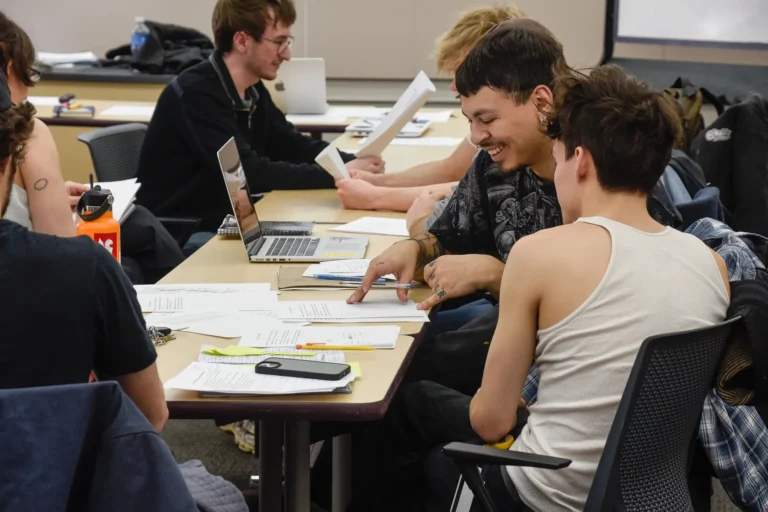 Two students lean over a shared desk in a classroom, talking and pointing at a set of printed papers while other classmates work in the background.