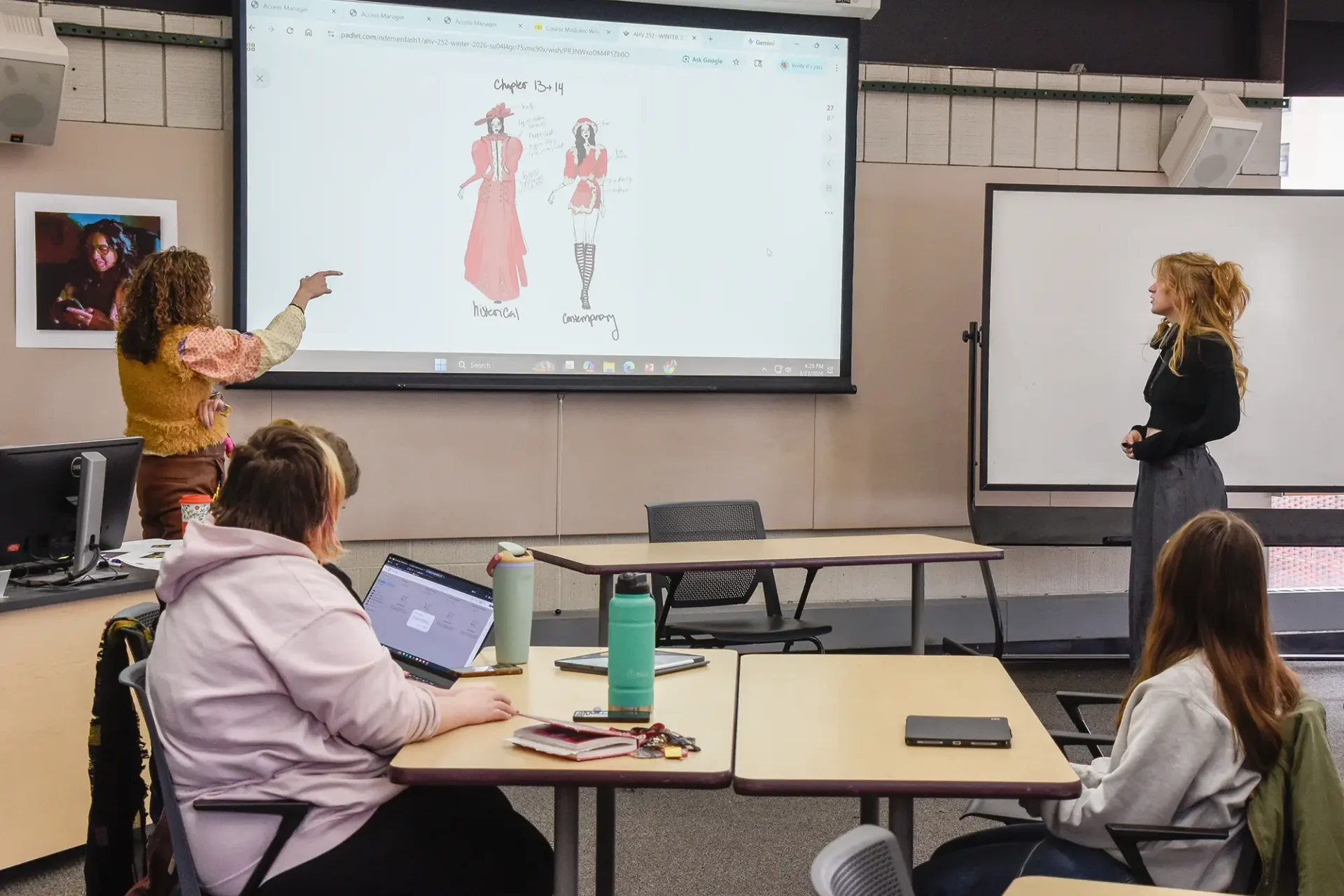 A classroom instructor points at a projector screen displaying two fashion sketches labeled "Historical" and "Contemporary." A student stands near a whiteboard at the front of the room, facing the instructor and classmates.